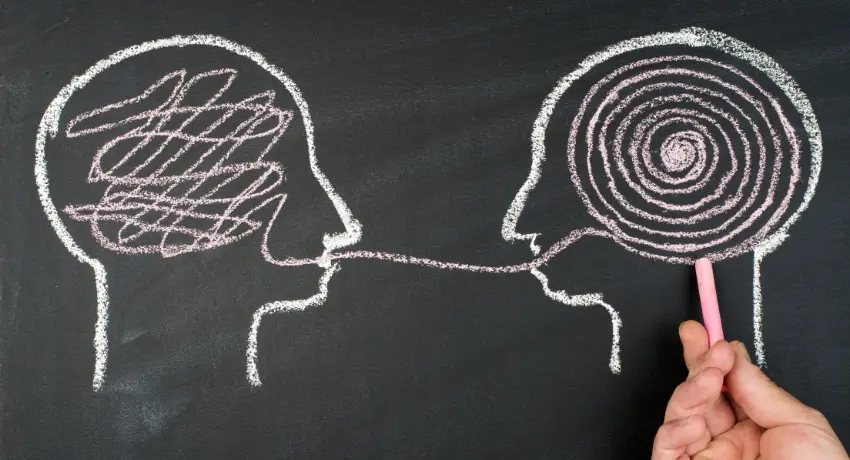 Desenho em giz em lousa preta de dois perfis de cabeça. Um cérebro tem riscos bagunçados e o outro tem um espiral, conectados por uma linha de comunicação. Mão segura o giz.