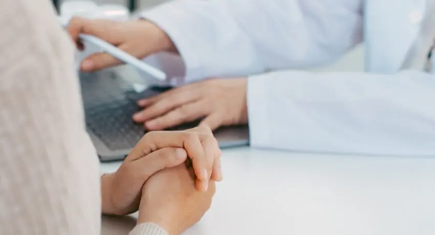 Paciente com mãos apoiadas na mesa conversa com profissional da saúde que utiliza notebook durante consulta em consultório, imagem usada em conteúdo sobre agência de marketing digital para saúde.